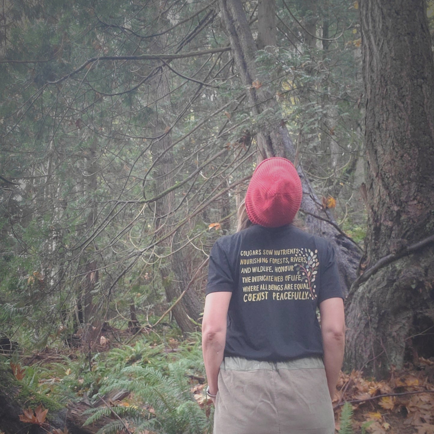 Person wearing a red beanie and dark shirt with text, standing in a forest.